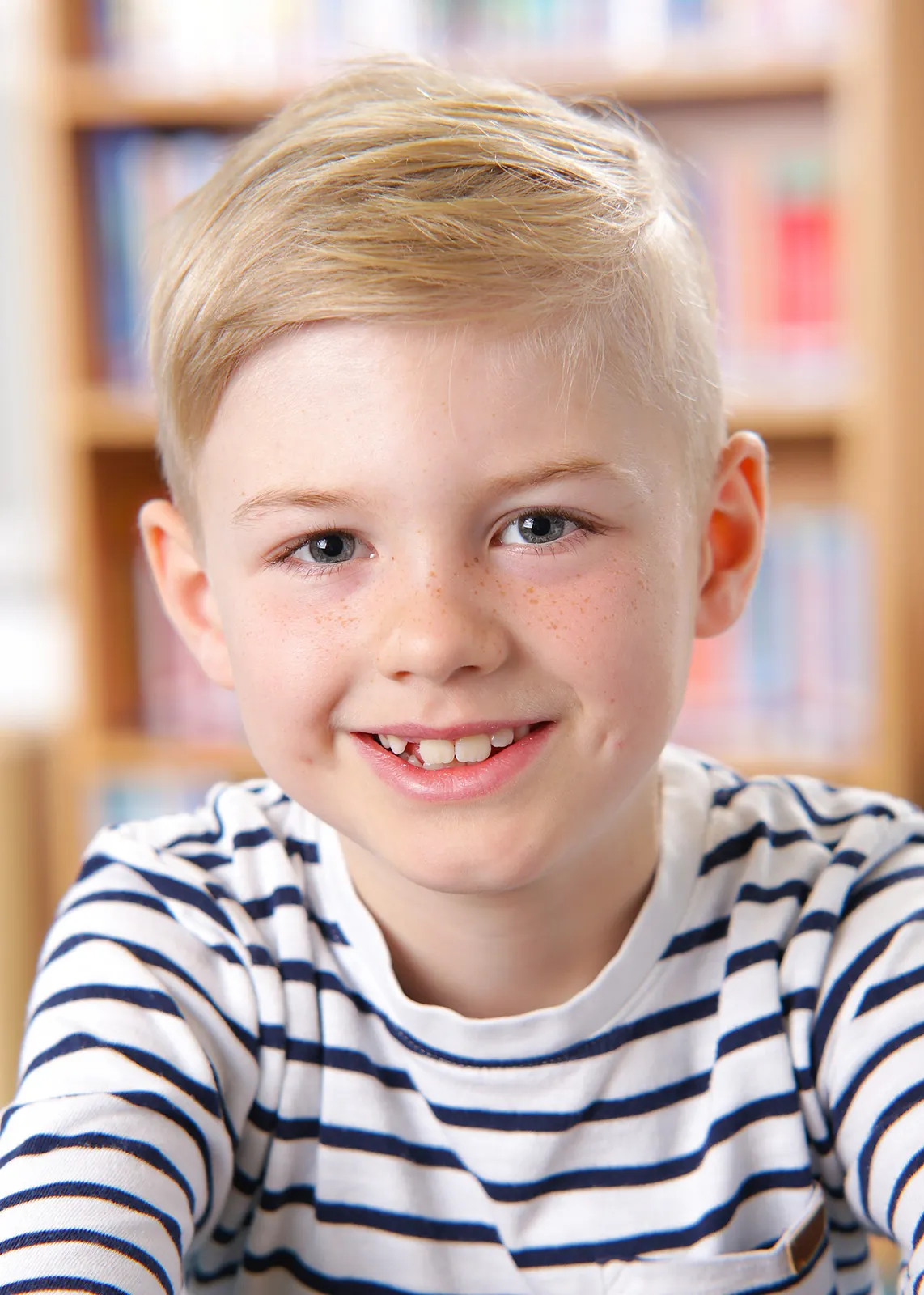 Blonder Erstklässler im gestreiften Pullover vor dem Bücherregal